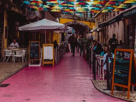 Pink street paving with cafe terrace tables and rainbow umbrellas above.