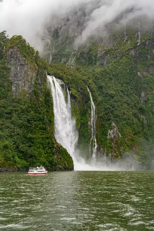 Waterfall cascading into Milford Sound fiord with small cruise boat.