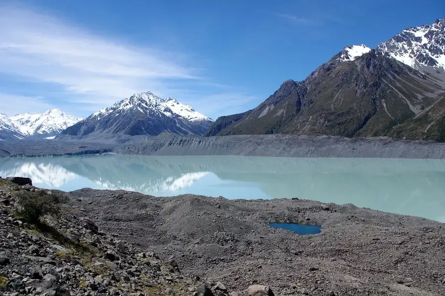 Grey volcanic terrain, Tasman glacier lake with snowy mountains behind.