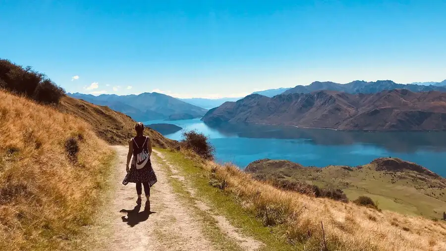 Woman walking on a track by a long lake beside mountains in sunshine..